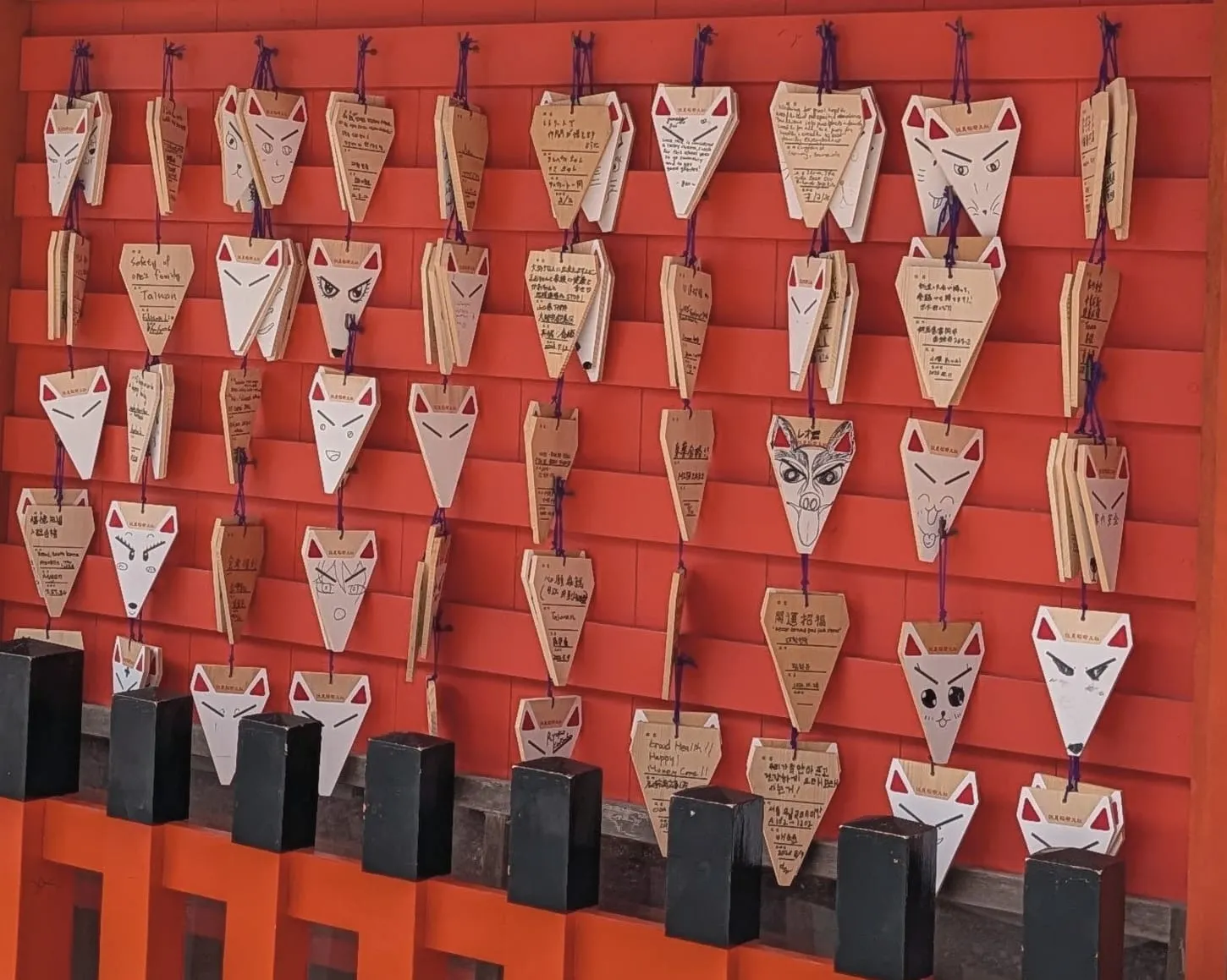 Fox-face ema with drawn-on faces hanging on a vermilion wall at Fushimi Inari
