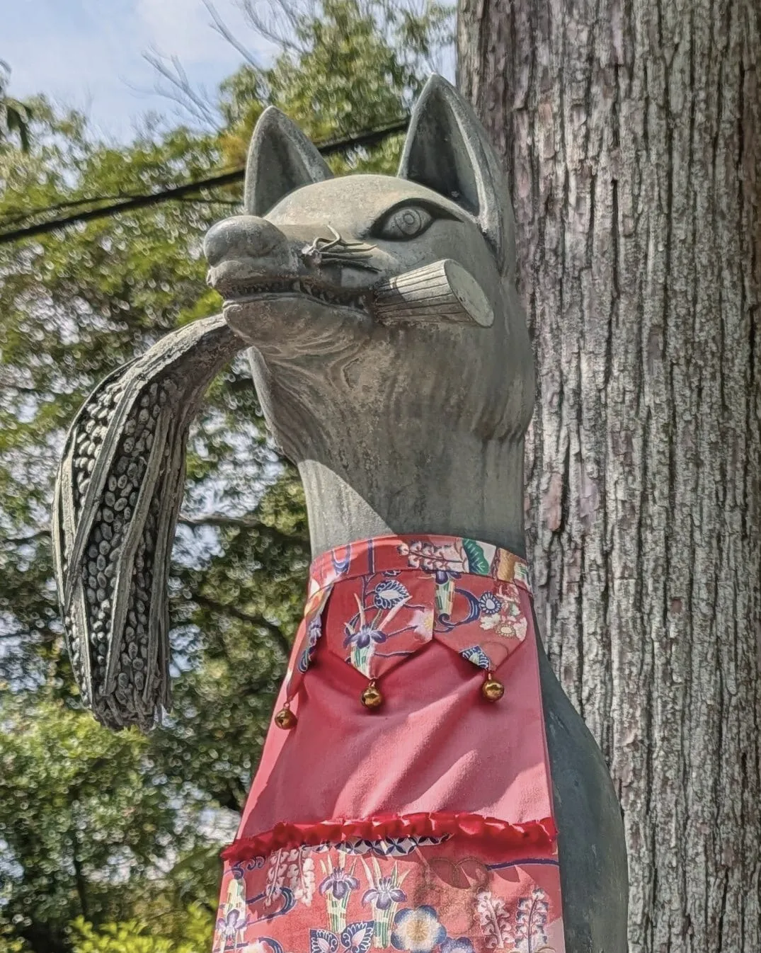 A bronze fox with a sheaf of rice in its mouth and a red bib with small bells, at Fushimi Inari, August 22, 2026