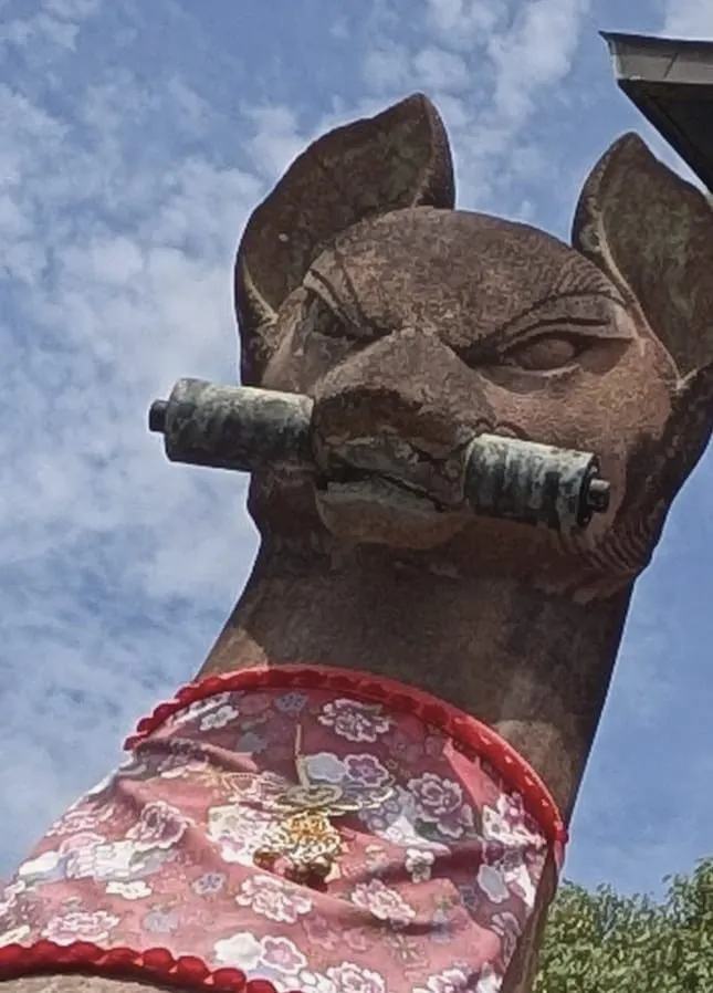 A stone fox with a scroll in its mouth and a flowered bib, at Fushimi Inari, August 22, 2026